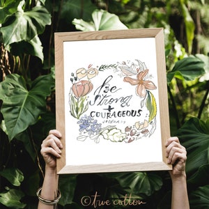 May include: A framed print with a floral wreath design and the text "Be strong + courageous Joshua 1:9" in black calligraphy. The print has a white background and is surrounded by a light brown frame. The text "true cotton" is written in a cursive font in the bottom right corner.