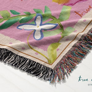 May include: A pink woven throw blanket with a floral design and the text "true cotton" in white script. The blanket has a fringe border with multicolored threads.