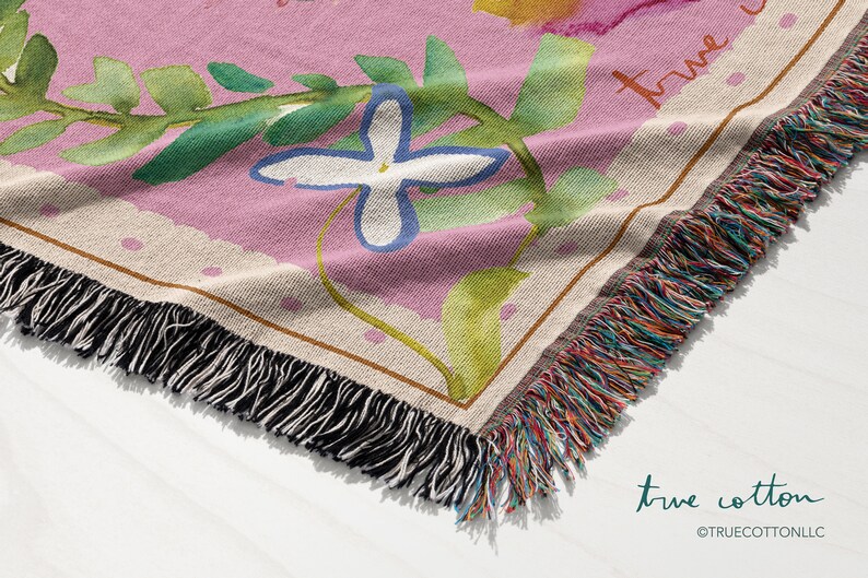 May include: A pink woven throw blanket with a floral design and the text "true cotton" in white script. The blanket has a fringe border with multicolored threads.