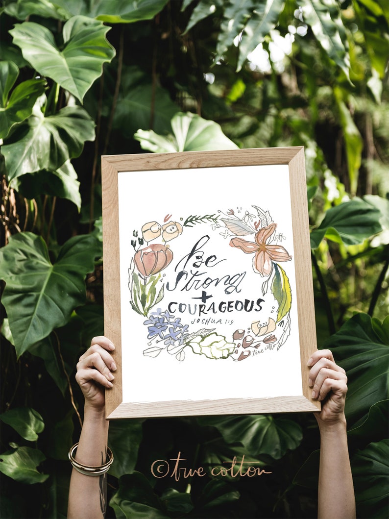 May include: A framed print with a floral wreath design and the text "Be strong + courageous Joshua 1:9" in black calligraphy. The print has a white background and is surrounded by a light brown frame. The text "true cotton" is written in a cursive font in the bottom right corner.