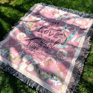 May include: A pink woven throw blanket with a floral pattern and the text "Let your unfailing love surround us Lord. For our hope is in you alone. Psalm 33:22"