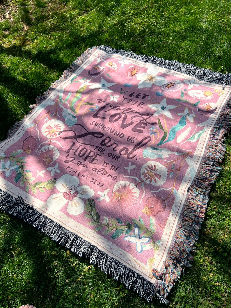 May include: A pink woven throw blanket with a floral pattern and the text "Let your unfailing love surround us Lord. For our hope is in you alone. Psalm 33:22"