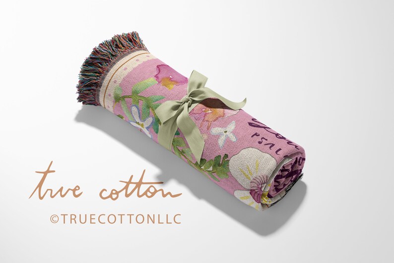 May include: A pink woven throw blanket with a floral pattern and the text "Just Breathe" in a script font. The blanket is rolled up and tied with a green ribbon.