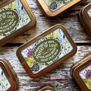 May include: Several rectangular, gold-colored tins with rounded edges are arranged on a wooden surface. Each tin features a floral design with text that reads "Purveyors of Hope, Keepers of Light, True Cotton, Our Daily Bread, Pocket Tin of Truth."