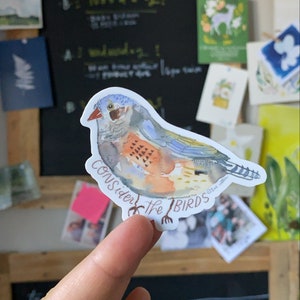 May include: A colorful bird sticker with the text "Consider the Birds." The sticker features a watercolor illustration of a bird with blue, orange, and brown feathers. The sticker is held up by a finger in front of a chalkboard.