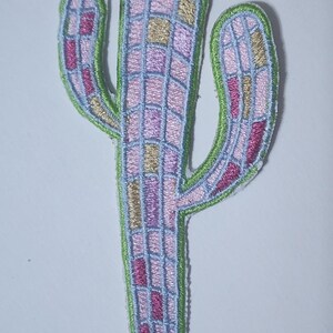 May include: Embroidered cactus patch in pink, purple, and gold hues, outlined in green. The cactus design features a grid pattern with light blue stitching. This decorative patch is ideal for adding a unique touch to clothing or accessories.