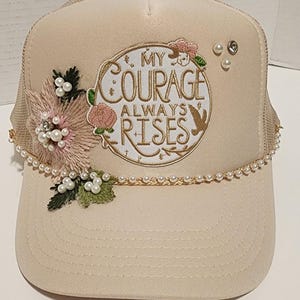 May include: A beige trucker hat with a mesh back and a curved brim. The front features an embroidered design with the words "My Courage Always Rises", floral accents, and faux pearls. A pearl strand trims the brim.