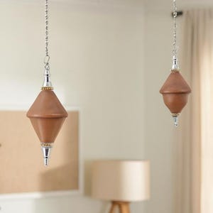 Può includere: Due tiranti per la luce marroni e argentati appesi al soffitto. Ogni tirante presenta un pendente marrone a forma di diamante con dettagli argentati e una catena. I pendenti sono in una stanza con una palette di colori neutri.