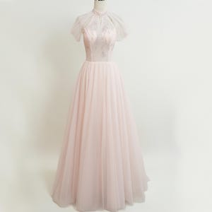 May include: A pale pink formal gown with a high neckline and short, sheer sleeves. The bodice features delicate lace detailing, and the skirt flows into a full, floor-length silhouette. The dress is displayed on a mannequin against a plain white background.