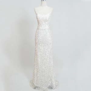 Puede incluir: Un vestido largo blanco con lentejuelas sobre un maniquí. El vestido presenta un corpiño ajustado con tirantes finos y una falda columna. Las lentejuelas crean un efecto brillante, reflejando la luz. El vestido se muestra sobre un fondo blanco.