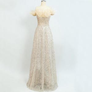 May include: A full-length, off-the-shoulder dress on a mannequin. The gown is a pale champagne colour and covered in sequins. The neckline and shoulders are trimmed with cream-coloured feathers. The dress features a fitted bodice and a flowing skirt.