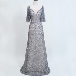 May include: A silver-grey, floor-length gown with a V-neck and short sleeves. The dress is covered in small, shimmering embellishments, creating a textured, sparkling effect. The dress is displayed on a mannequin against a plain white background.