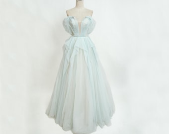 May include: A light blue off-the-shoulder gown with a plunging neckline and a flowing skirt. The bodice features delicate embellishments, and the dress is displayed on a mannequin. The gown's design includes layered ruffles and a soft, ethereal appearance.