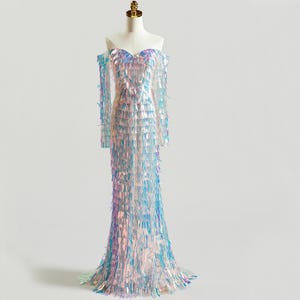 Puede incluir: Vestido largo con hombros descubiertos sobre un maniquí. El vestido está cubierto de lentejuelas iridiscentes y con flecos en tonos azules, rosas y plateados. El corpiño tiene un escote corazón y las mangas son largas y con flecos.
