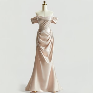 May include: A champagne-colored, off-the-shoulder formal gown. The dress features a draped bodice and a flowing, floor-length skirt. The dress is displayed on a mannequin against a plain white background.