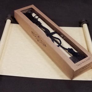 May include: A cream-colored paper scroll with dark brown wooden finials at each end, partially unrolled. A rectangular brown cardboard box with a clear window rests on top, containing a black braided cord and the text Say it on a Scroll 8.5 x 11.