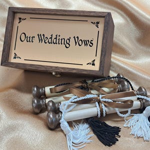 May include: A wooden box with the text "Our Wedding Vows" on a gold background. Below the box are several rolled scrolls tied with white and brown ribbons, and tassels. The scrolls have decorative metal ends.