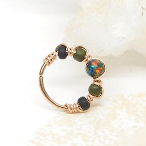 May include: A circular hoop ring crafted from thin rose gold wire, featuring a central iridescent dark opal bead flanked by two smaller olive green beads and two matte black beads, all secured with delicate wire wrapping techniques.