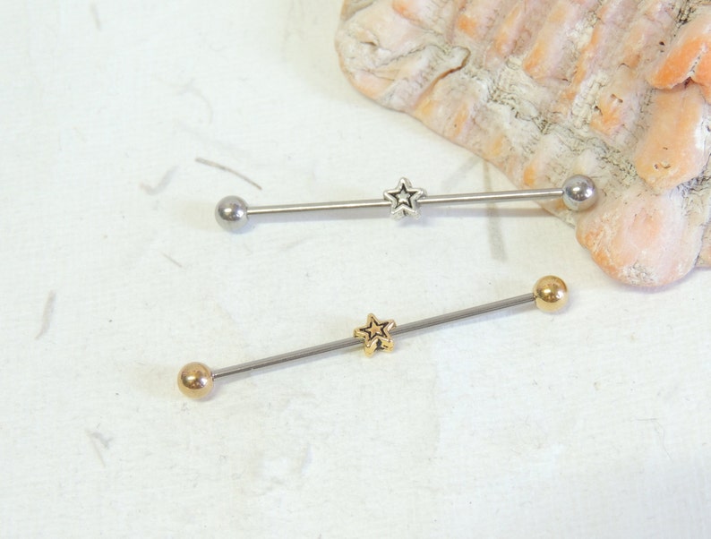 May include: Two silver barbell body jewelry pieces with star-shaped charms. The top barbell has a silver charm and the bottom barbell has a gold charm.