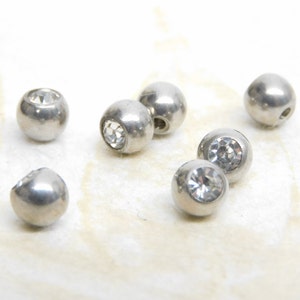 May include: Six silver-toned metal balls with clear rhinestones in the centre. The balls are for body jewellery.