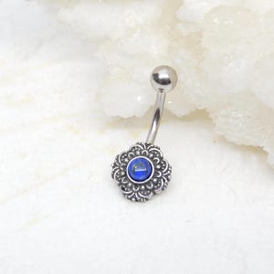 May include: A silver belly button ring with a blue gemstone set in a decorative floral design.