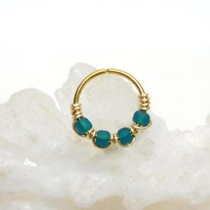 May include: A gold-colored nose ring featuring five teal-colored beads. The beads are evenly spaced around the ring and are secured with gold-colored wire. The ring is set against a white, textured background.