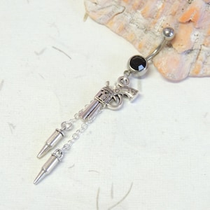 May include: Silver-toned belly button ring featuring a revolver charm with dangling bullet pendants. The ring has a curved barbell design with a black gem and a small silver ball at the top.