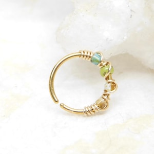 May include: A gold-colored nose ring with a small opening. The ring is adorned with small, colorful beads in shades of green, blue, and tan, wrapped with gold wire. The jewelry is set against a white background.