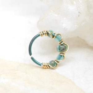 May include: A teal nose ring with gold wire accents and small blue-green beads. The ring is a circular shape, with the beads and wire wrapped around the top portion. The metal has a slightly textured appearance.