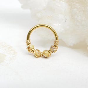 May include: A gold-colored septum ring with small, light-colored beads wrapped in gold wire. The ring is circular and has a hinged closure. The beads have a speckled appearance. The jewelry is set against a white background.