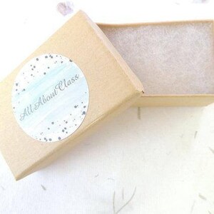 Puede incluir: Una peque&ntilde;a caja de cart&oacute;n marr&oacute;n con una pegatina blanca moteada que dice "All About Glass". La caja est&aacute; abierta y el interior est&aacute; forrado con papel de seda blanco.