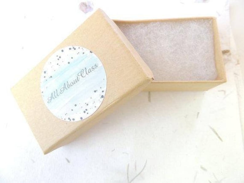 Puede incluir: Una peque&ntilde;a caja de cart&oacute;n marr&oacute;n con una pegatina blanca moteada que dice "All About Glass". La caja est&aacute; abierta y el interior est&aacute; forrado con papel de seda blanco.
