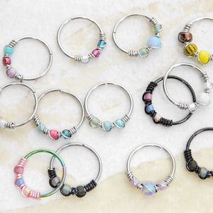 May include: A collection of 12 silver and black hoop earrings with various coloured beads and charms. The earrings are all different designs and feature a variety of colours, including pink, blue, green, yellow, and white.