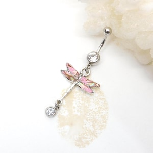 May include: A silver-colored belly button ring featuring a dragonfly design. The dragonfly has pink and iridescent wings and a clear gem at the bottom. The top and middle of the ring also have clear gems.