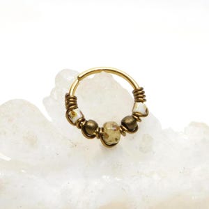 May include: A gold-colored nose ring with a hinged clasp. The ring is adorned with small, round, and oblong beads in shades of brown, cream, and gold. The beads are wire-wrapped around the ring, creating a unique, handcrafted look. The jewelry is set against a white background.