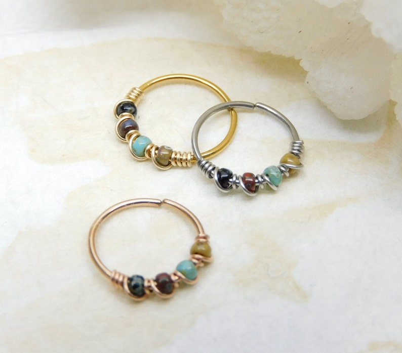 May include: Three nose rings with colourful beads. The rings are made of gold, silver, and rose gold. The beads are turquoise, brown, and black.