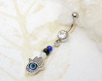 May include: A silver belly button ring with a dangling charm. The charm features a silver Hamsa hand with a blue eye, and is accented with black, blue, and white beads. The top of the ring has a clear gem.