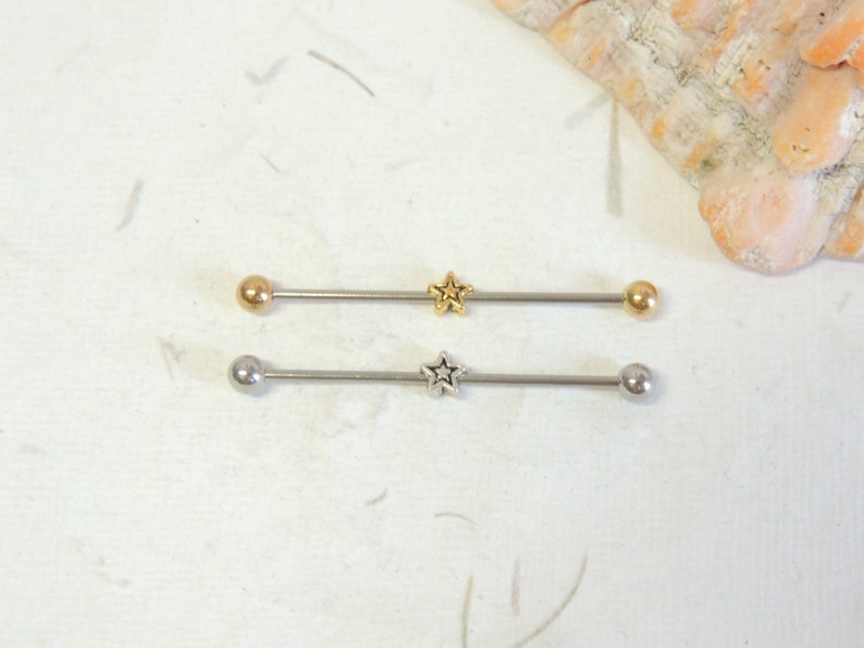 May include: Two surgical steel barbell body jewelry pieces with star-shaped charms. The top barbell is gold-toned and the bottom barbell is silver-toned.