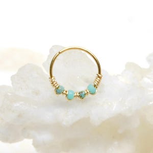 May include: A gold-colored nose ring with small, light blue beads. The beads are evenly spaced along the bottom of the ring. The ends of the ring are wrapped with gold-colored wire.