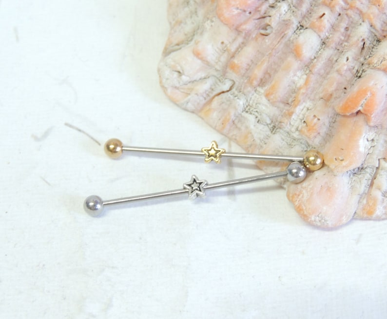 May include: Two silver barbell style body jewelry pieces with gold star accents. The jewelry is on a white background.