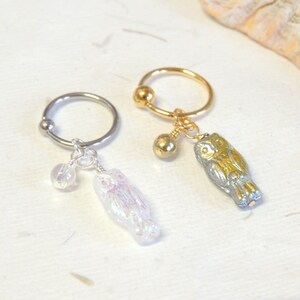 May include: Two silver hoop earrings with a small charm attached to each. One charm is a silver owl with iridescent white wings and a clear crystal bead. The other charm is a gold owl with iridescent green wings and a gold bead.