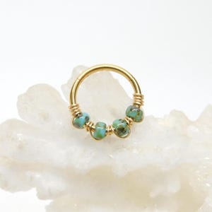 May include: A gold-colored hoop earring with five turquoise-colored beads. The beads are spaced evenly around the hoop, and the wire is wrapped around the hoop on either side of the beads. The earring is on a white surface.