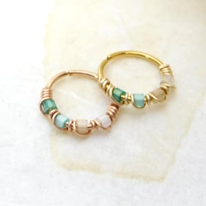 May include: Two hoop earrings. One is gold-colored, the other is copper-colored. Both are adorned with small, colorful beads in shades of green, blue, and beige, wrapped with wire. The earrings are designed for pierced ears.