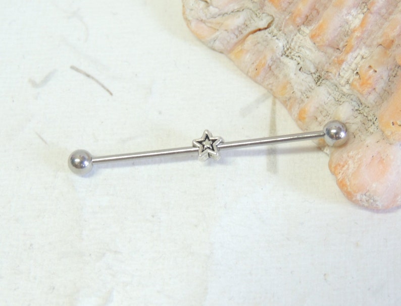 May include: Silver barbell with a star charm in the center. The barbell is a body piercing jewelry item.
