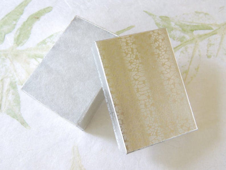Puede incluir: Dos cajas de regalo rectangulares. Una caja es plateada con una superficie texturizada, y la otra es dorada con un patr&oacute;n craquelado. Las cajas est&aacute;n ligeramente anguladas, mostrando su brillo met&aacute;lico. El fondo es un blanco roto suave con sutiles acentos de hojas verdes.