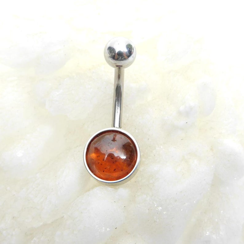 Puede incluir: Un piercing de ombligo plateado con una piedra redonda de &aacute;mbar naranja.