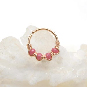 May include: A gold-colored nose ring with five pink beads. The beads are evenly spaced along the bottom of the ring. The ring is made of a thin wire that is wrapped around the beads and the ring itself.