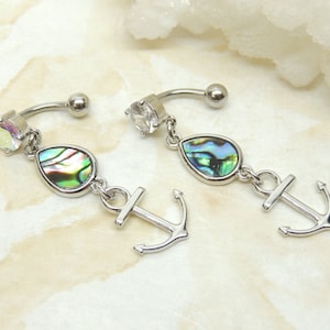 May include: A pair of silver belly button rings with a teardrop-shaped abalone shell charm and an anchor charm. The rings are curved and have a clear crystal on the top.