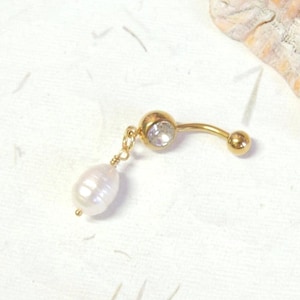 May include: A gold-toned curved barbell belly ring featuring a clear round gemstone at the top and a dangling white oval-shaped freshwater pearl attached by a small gold-toned wire loop. The jewelry piece has a polished metallic finish throughout.
