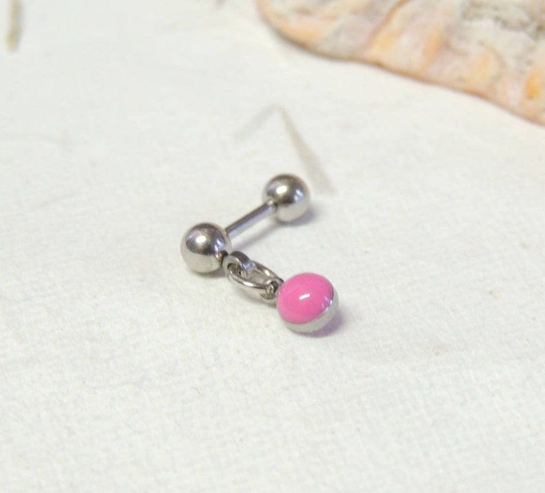 May include: A silver barbell earring with a dangling pink charm. The earring has a straight post with a ball at each end. The charm is a small, round pink bead with a silver border, attached by a small ring.